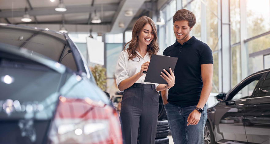 Two people standing in a car dealership, with one holding a tablet and explaining something to the other