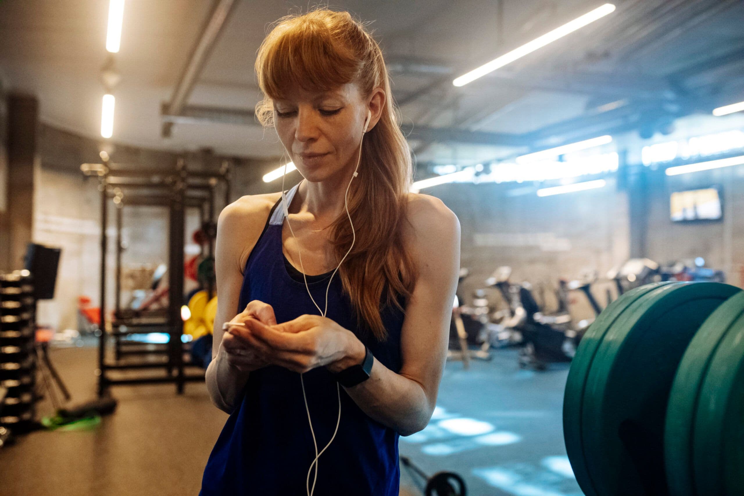 Person in a gym wearing a blue tank top and earphones, holding a smartphone. Background includes gym equipment like weight racks and exercise machines