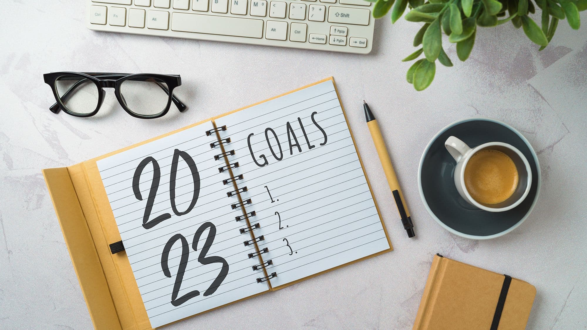 Notebook with text "2023 goals", lying on a table next to cup with coffee, glasses, pencil, computer keyboard and another notebook
