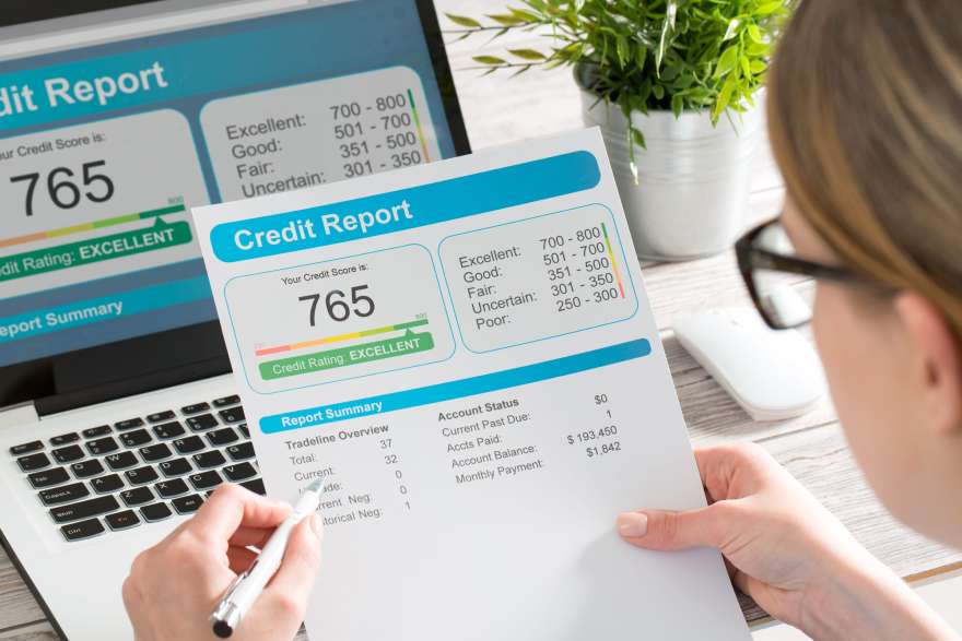 Person reviewing a printed credit report while also viewing the same report on a laptop screen. Credit score shown is 765 with an 'Excellent' rating. Report includes account balances, payment history, and credit rating scale