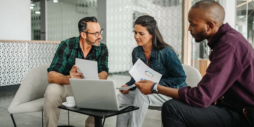 Two men and one woman sitting on chairs in front of computer, keeping papers with charts in their hands