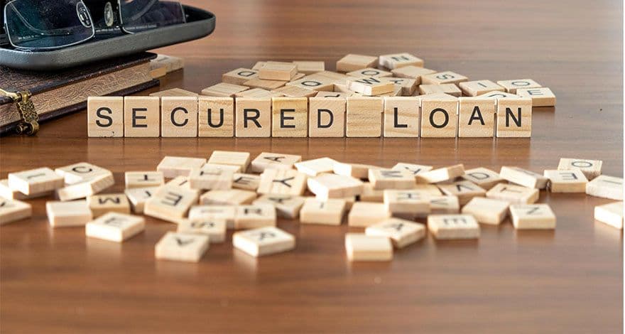 Wooden letter tiles spelling out 'SECURED LOAN' on a wooden surface, with scattered tiles, a pair of glasses, and a notebook in the background