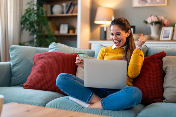 Person sitting on a couch with red cushions, wearing a yellow sweater and blue jeans, holding a credit card and using a laptop